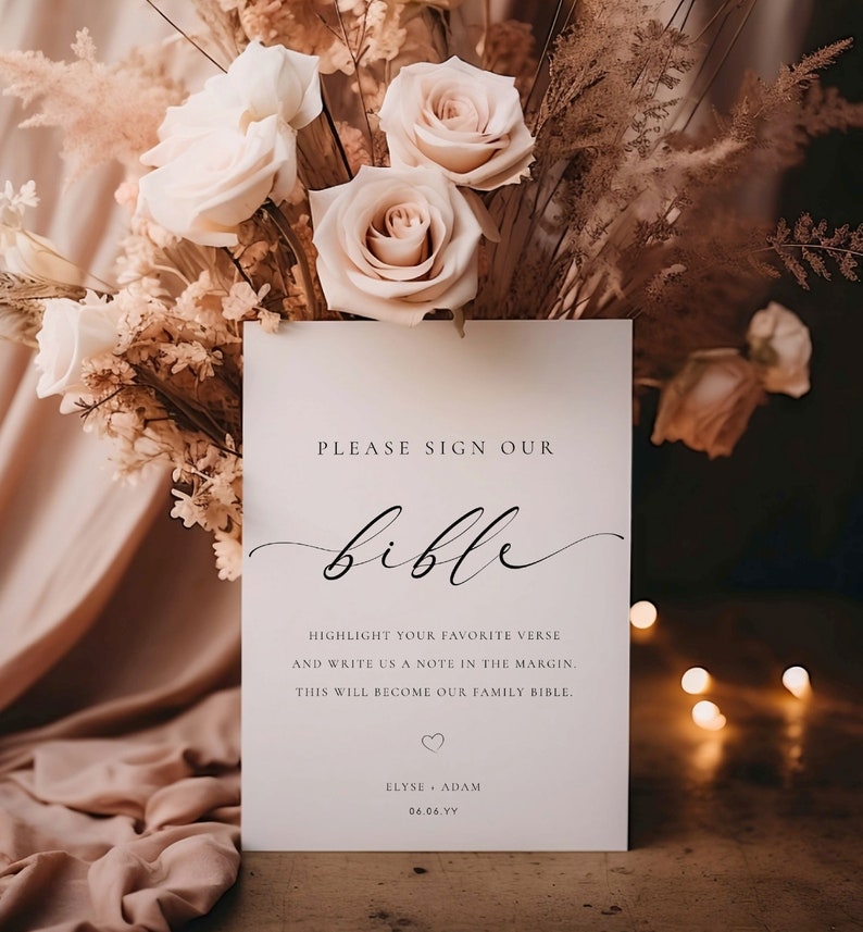 May include: A white sign with black text that reads "Please sign our bible. Highlight your favorite verse and write us a note in the margin. This will become our family bible. Elyse Adam 06.06.YY". The sign is sitting on a wooden table with a bouquet of beige roses and dried grasses in the background.