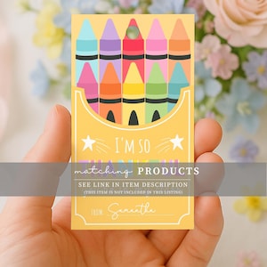 May include: A yellow card featuring a crayon box design with colorful crayons. The card has the text "I'M SO" and "matching PRODUCTS". The card is held in a hand, with a floral background.