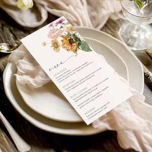 May include: A white menu card with a floral design featuring sunflowers and other flowers. The menu card lists appetizers, starters, main course, and dessert options. The text on the menu card reads: "meru", "APPETIZER", "Antipasto Platters", "Local charcuterie, farmhouse cheeses, quince paste, lavosh, fresh and dried fruit.", "STARTERS", "Farmhouse fresh greens garden salad served with raspberry infused vinaigrette.", "MAIN COURSE", "Twice-cooked shoulder of lamb with roasted vegetables and red wine jus.", "Honey chicken, crispy speck, steamed greens and crispy potatoes.", "DESSERT", "Tahition Vanilla sponge with homemade lemon curd."