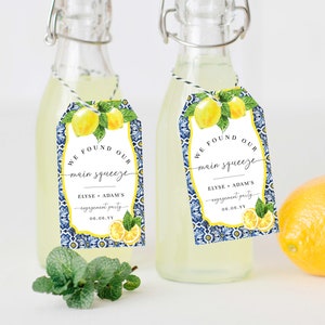 Può includere: Due piccole bottiglie di vetro riempite con un liquido trasparente, ciascuna con un'etichetta a motivi blu e bianchi che dice "We found our main squeeze Elyse + Adam's engagement party 06.06.YY". Le etichette raffigurano limoni e foglie verdi.