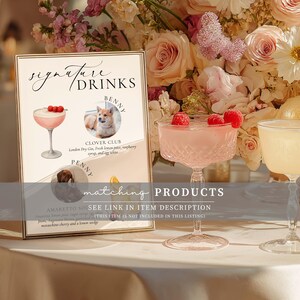 May include: A framed sign with "Signature Drinks" text, featuring illustrations of cocktails and pet portraits. A pink cocktail with raspberries sits in a crystal glass. Floral arrangements and a second cocktail glass are in the background.