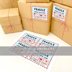 Peut inclure: Plusieurs paquets emball&eacute;s dans du papier brun, attach&eacute;s avec de la ficelle rouge et blanche. Chaque paquet porte une &eacute;tiquette bleue et blanche "Fragile, Manipuler avec soin" avec des illustrations festives et le texte "Cadeau du P&egrave;re No&euml;l."