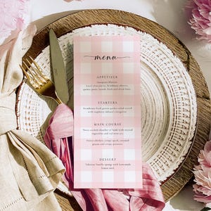 Puede incluir: Una tarjeta de menú de vichy rosa y blanco con la palabra "menú" escrita en cursiva negra. El menú enumera aperitivos, entrantes, platos principales y postres. El menú está en un plato de madera blanco y marrón con una servilleta rosa.