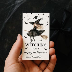 Op de afbeelding: Een rechthoekig Halloween-cadeaulabel met een heks op een bezemsteel, omringd door vleermuizen en sterren. Het label leest "Witching You a Happy Halloween" en bevat ruimte voor een naam.