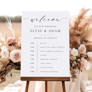 May include: A white wedding sign on a wooden easel with the text "welcome" in elegant script. The sign lists the order of events for Elyse & Adam's wedding, including times for the ceremony, dinner, and first dance. Floral arrangements frame the sign.