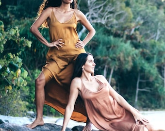 May include: Two women wearing flowing, silk-like dresses pose on a large rock near a beach. One dress is a golden-brown color, and the other is a muted rose color. The dresses have a simple, spaghetti-strap design.