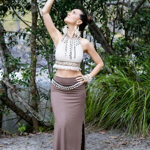 K&ouml;nnte beinhalten: Ein cremefarbenes Neckholder-Bauchfrei-Top mit braunem Mustersaum und Fransen, kombiniert mit einem langen braunen Rock mit Seitenschlitz und einem Tailleng&uuml;rtel aus wei&szlig;en Kaurimuscheln. Das Outfit umfasst eine passende Kaurimuschel-Halskette und ein Armband.