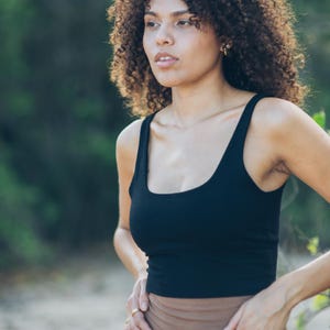 Könnte beinhalten: Ein schwarzes Tanktop mit einem Rundhalsausschnitt. Das Oberteil ist aus einem weichen, dehnbaren Material gefertigt. Das Model trägt einen goldenen Ring und Ohrringe. Der Hintergrund ist ein verschwommenes Grün.