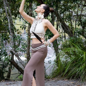K&ouml;nnte beinhalten: Ein cremefarbenes Neckholder-Oberteil mit Makramee-Details und Fransen, kombiniert mit einem einfarbigen braunen Maxirock mit hohem Seitenschlitz. Das Outfit wird durch eine Kauri-Muschel-Halskette und einen passenden Muschelg&uuml;rtel erg&auml;nzt.