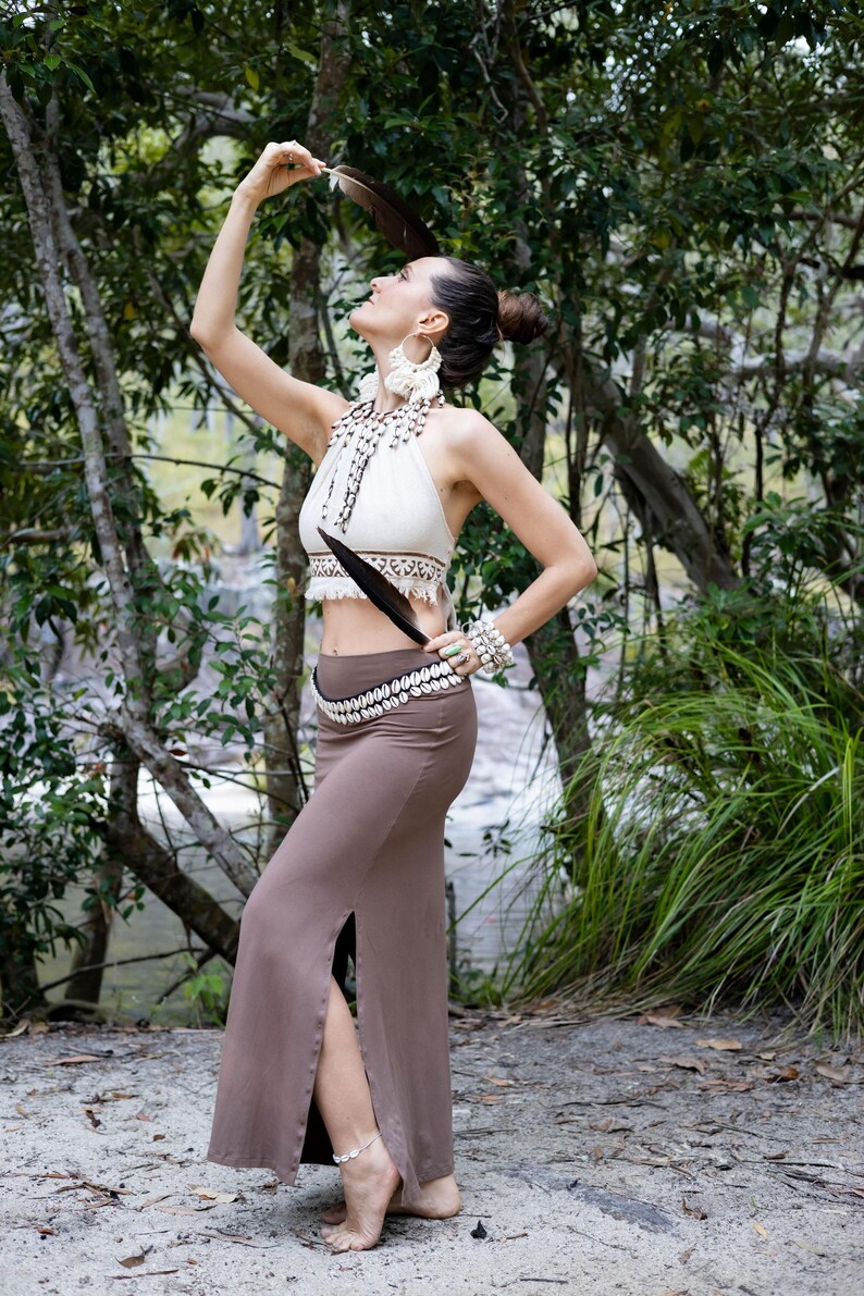K&ouml;nnte beinhalten: Ein cremefarbenes Neckholder-Oberteil mit Makramee-Details und Fransen, kombiniert mit einem einfarbigen braunen Maxirock mit hohem Seitenschlitz. Das Outfit wird durch eine Kauri-Muschel-Halskette und einen passenden Muschelg&uuml;rtel erg&auml;nzt.