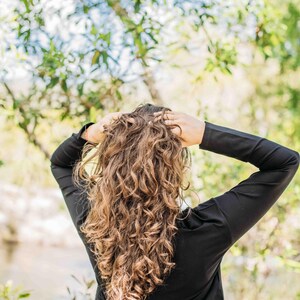 May include: A person with long, curly brown hair, wearing a black long-sleeved top and olive green pants, stands with their back to the camera. The person's hands are in their hair. The background is a blurred natural setting.