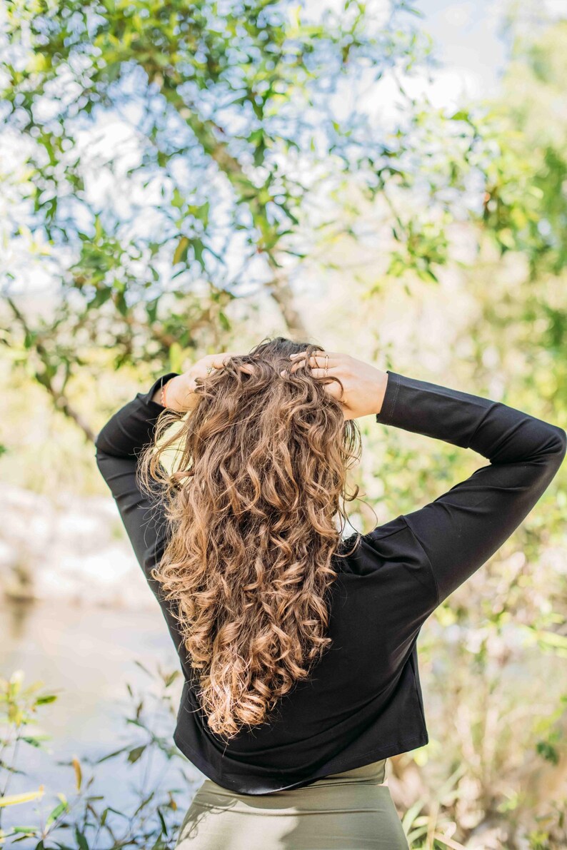 May include: A person with long, curly brown hair, wearing a black long-sleeved top and olive green pants, stands with their back to the camera. The person's hands are in their hair. The background is a blurred natural setting.