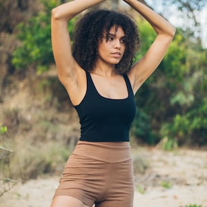 Pu&ograve; includere: Un top nero e pantaloncini color beige. Il top ha una scollatura quadrata e i pantaloncini sono a vita alta. La persona &egrave; all'aperto con le braccia alzate in una posa yoga. Lo sfondo &egrave; sfocato con alberi e fogliame.
