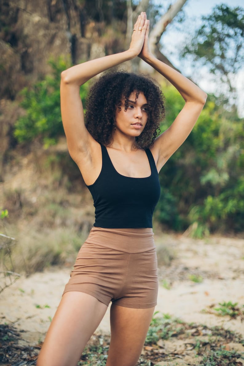 Pu&ograve; includere: Un top nero e pantaloncini color beige. Il top ha una scollatura quadrata e i pantaloncini sono a vita alta. La persona &egrave; all'aperto con le braccia alzate in una posa yoga. Lo sfondo &egrave; sfocato con alberi e fogliame.