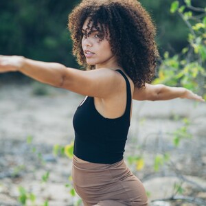 Pu&ograve; includere: Una donna in posa yoga, indossa un top senza maniche nero e pantaloncini marroni. La donna ha i capelli castani ricci e si trova all'aperto con uno sfondo verde sfocato.