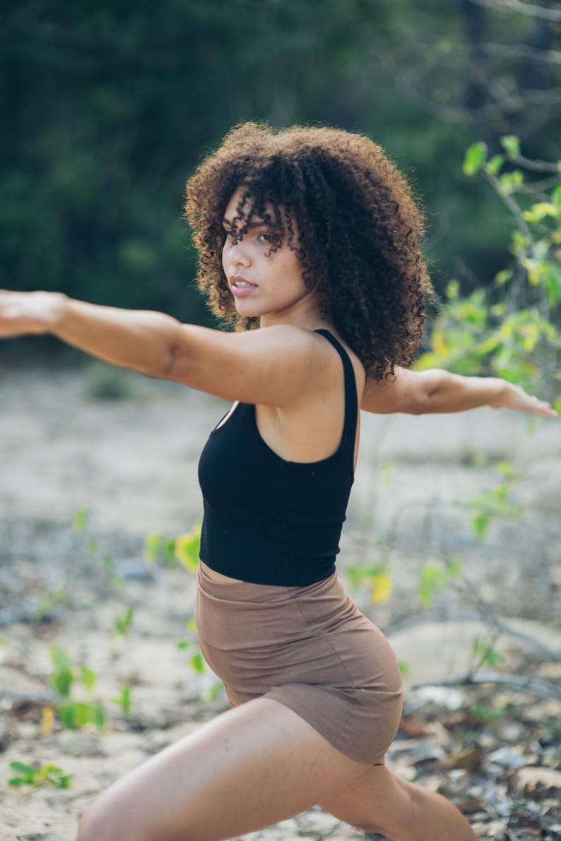 Pu&ograve; includere: Una donna in posa yoga, indossa un top senza maniche nero e pantaloncini marroni. La donna ha i capelli castani ricci e si trova all'aperto con uno sfondo verde sfocato.