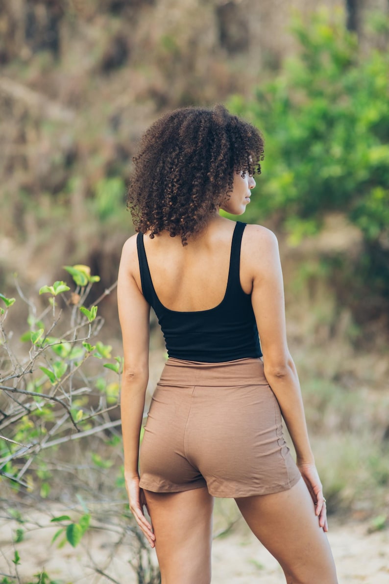 Pu&ograve; includere: Una donna indossa una canotta nera e pantaloncini marroni. I pantaloncini sono a vita alta e dal design semplice. Lo sfondo &egrave; sfocato, con fogliame verde in un ambiente naturale.