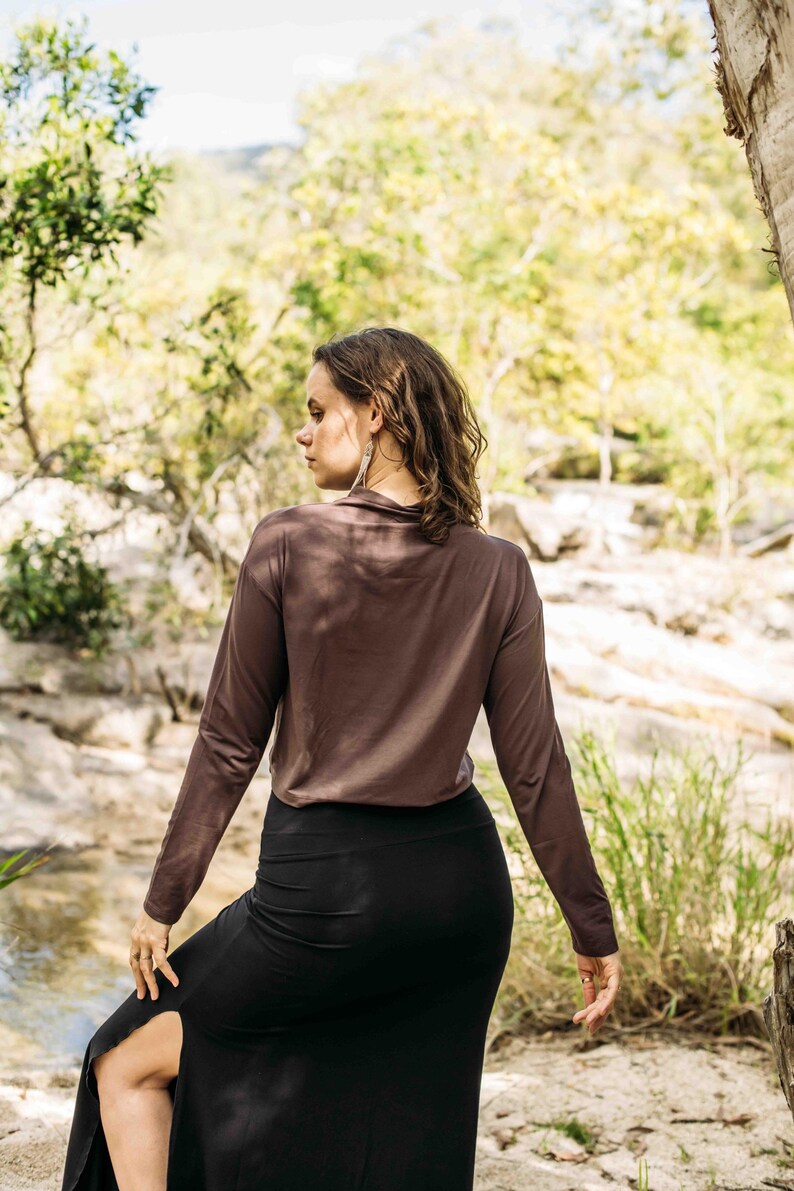 May include: A woman wearing a long-sleeved brown top and a black skirt with a high slit. The outfit is simple and elegant, suitable for casual wear. The background features a natural outdoor setting with trees and a stream.