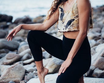 Può includere: Un top corto con un motivo beige e nero abbinato a leggings neri. La modella è inginocchiata su una roccia vicino all'oceano. Il top ha spalline sottili e un orlo grezzo.