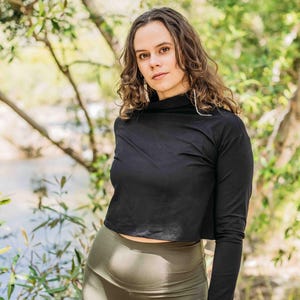 May include: A black long-sleeved crop top with a high neckline paired with olive green leggings. The model is standing outdoors with a natural background of trees and a body of water.