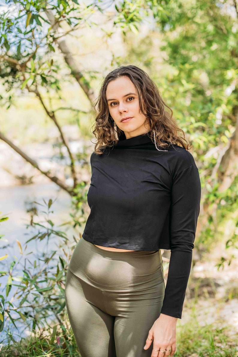 May include: A black long-sleeved crop top with a high neckline paired with olive green leggings. The model is standing outdoors with a natural background of trees and a body of water.