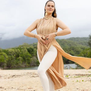Op de afbeelding: Een vrouw draagt een vloeiende, bruin-beige wikkeljurk over een witte legging, staand op een zandstrand. De jurk heeft lange, draperende panelen. Accessoires zijn gouden sieraden. De achtergrond toont een groene boomgrens en een bewolkte lucht.