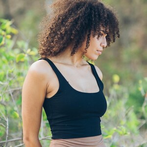 Pu&ograve; includere: Un top nero con scollo tondo e spalline larghe. Il top &egrave; abbinato a leggings a vita alta color beige. La modella ha i capelli castani ricci e si trova all'aperto con un fogliame verde sullo sfondo.