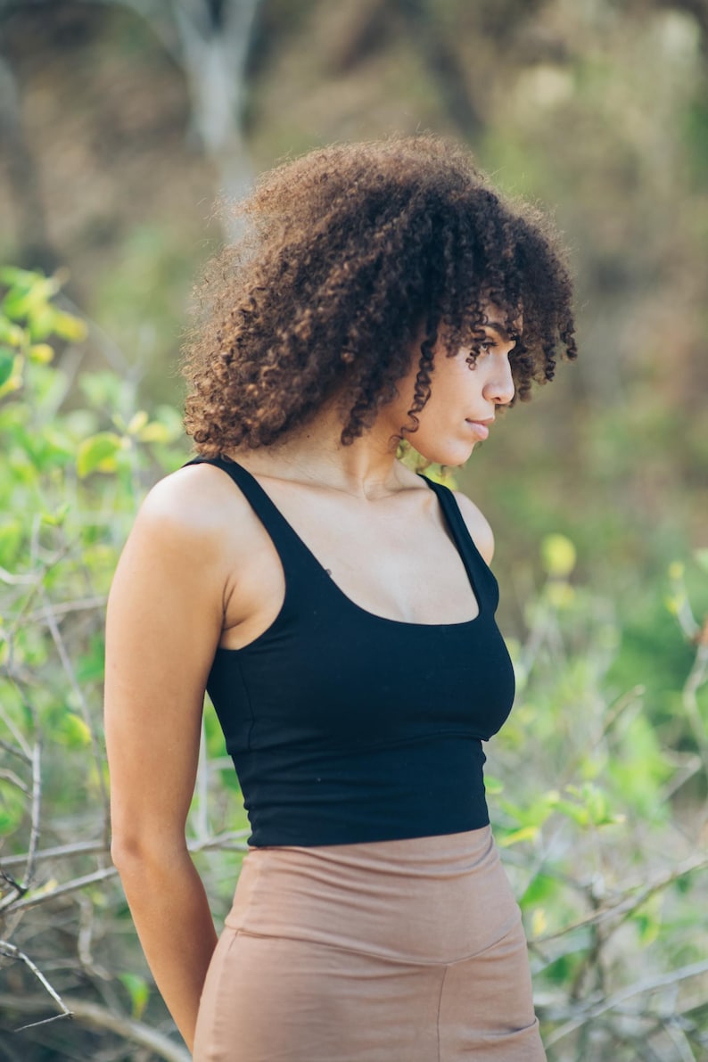Pu&ograve; includere: Un top nero con scollo tondo e spalline larghe. Il top &egrave; abbinato a leggings a vita alta color beige. La modella ha i capelli castani ricci e si trova all'aperto con un fogliame verde sullo sfondo.