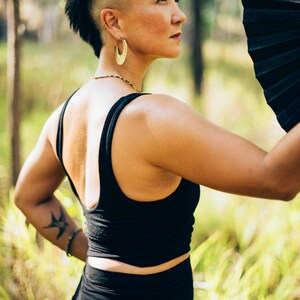 Pu&ograve; includere: Una persona con canotta e gonna nere, che tiene in mano un ventaglio nero. La persona ha un'acconciatura mohawk e orecchini a cerchio dorati. Un tatuaggio &egrave; visibile sul braccio sinistro. Lo sfondo &egrave; una scena di foresta sfocata.