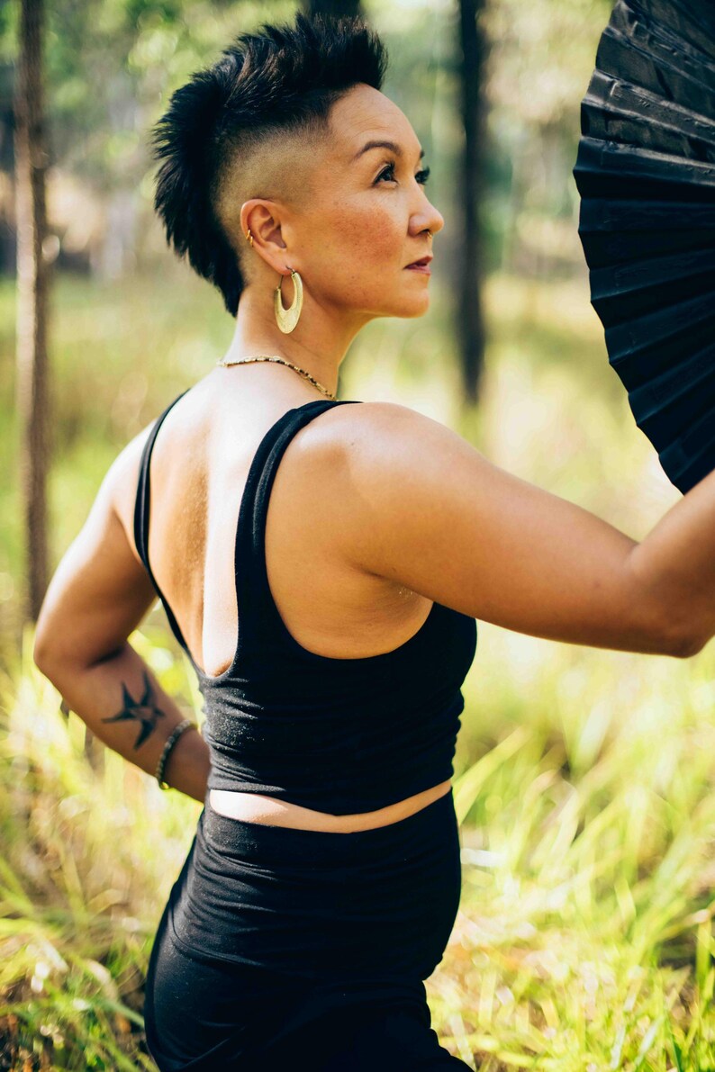 Pu&ograve; includere: Una persona con canotta e gonna nere, che tiene in mano un ventaglio nero. La persona ha un'acconciatura mohawk e orecchini a cerchio dorati. Un tatuaggio &egrave; visibile sul braccio sinistro. Lo sfondo &egrave; una scena di foresta sfocata.