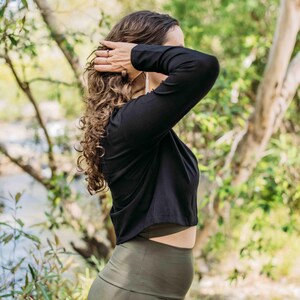 May include: A black long-sleeve crop top and olive green leggings. The model is outdoors with curly hair, and is touching their hair. The background is a blurred view of trees and a river.
