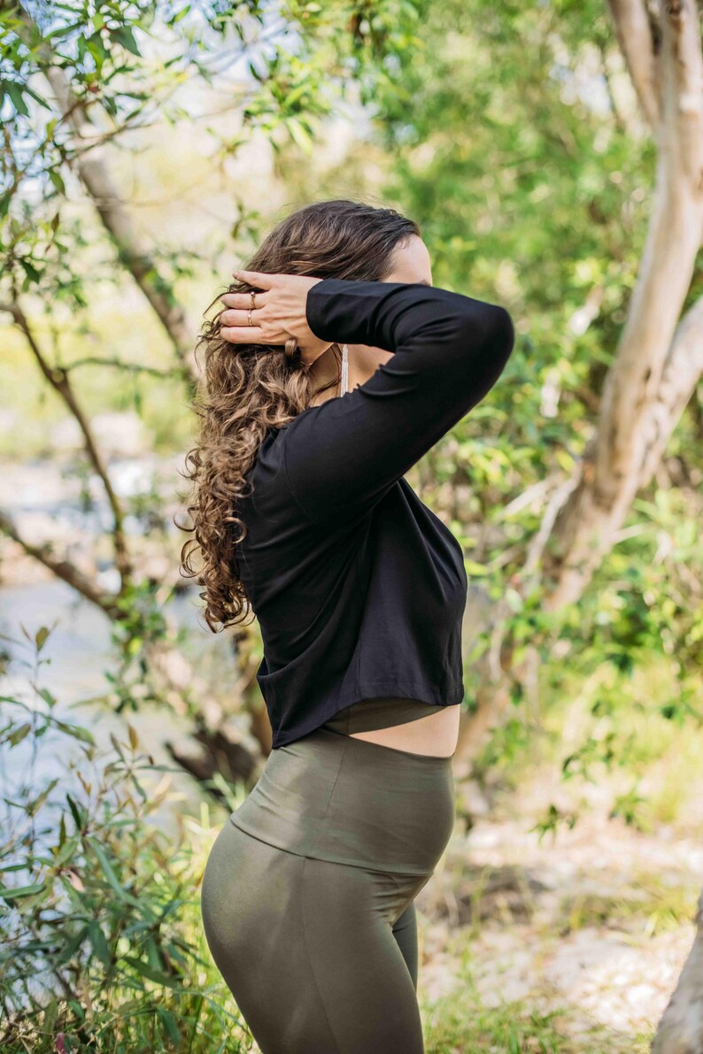 May include: A black long-sleeve crop top and olive green leggings. The model is outdoors with curly hair, and is touching their hair. The background is a blurred view of trees and a river.