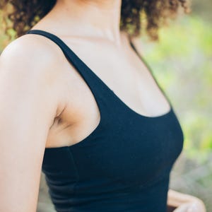 Pu&ograve; includere: Un top nero con scollo tondo. La parte superiore &egrave; realizzata in un materiale morbido ed elastico. La modella indossa orecchini a cerchio dorati. Lo sfondo &egrave; sfocato con toni verdi e marroni.