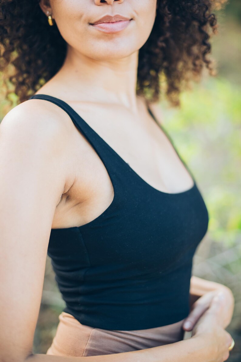 Pu&ograve; includere: Un top nero con scollo tondo. La parte superiore &egrave; realizzata in un materiale morbido ed elastico. La modella indossa orecchini a cerchio dorati. Lo sfondo &egrave; sfocato con toni verdi e marroni.