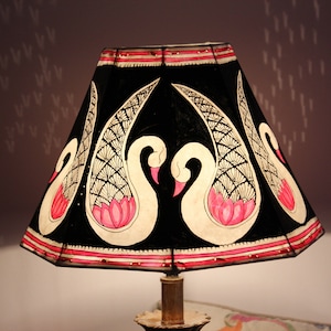 May include: A black lampshade with a hand-painted design featuring white swans with pink accents and a pink lotus flower. The lampshade has a red trim and is set on a brass lamp base. The overall theme is decorative lighting.