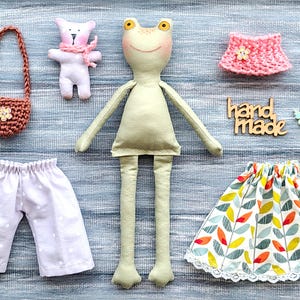 May include: A handmade frog doll with long limbs, surrounded by miniature clothing items. The set includes a crocheted purse, a bear, a pink skirt, a pair of polka-dot pants, and a leaf-patterned skirt. The word "handmade" is also visible.