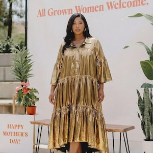 May include: A woman wearing a gold metallic midi dress with tiered ruffles and bell sleeves. She is wearing red heels. The background includes a sign that reads "All Grown Women Welcome" and "Happy Mother's Day!"