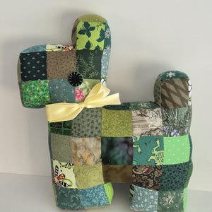 May include: A handmade dog-shaped pillow crafted from patchwork fabric in various shades of green, brown, and blue. The pillow features a yellow ribbon and a black button. This decorative item is suitable for home decor.