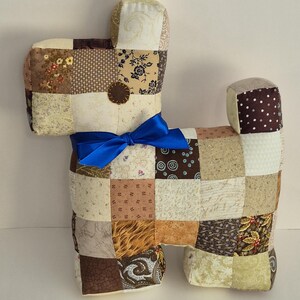 May include: A patchwork dog-shaped pillow in various neutral tones, including beige, brown, and cream, with a blue ribbon tied around its neck. The pillow is made of fabric squares and has a small, round, brown button detail.