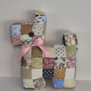 May include: A patchwork dog-shaped stuffed animal with a pink ribbon tied around its neck. The dog is made of various fabric squares in floral and geometric patterns, including red, blue, and brown. The dog's ears and tail are also made of patchwork fabric.