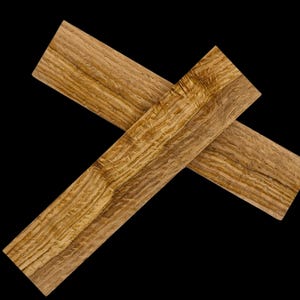 Puede incluir: Dos tablas de madera, cruzadas en forma de X, sobre un fondo negro. Las tablas, de color marrón claro, presentan vetas de madera visibles. La madera parece recién cortada, ideal para un proyecto de carpintería.
