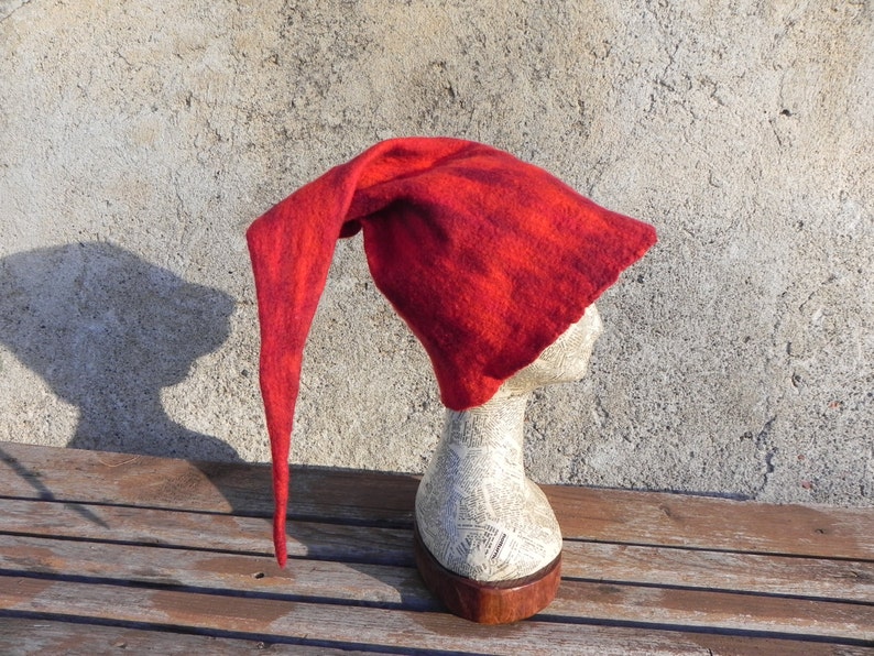 May include: A red felt hat with a long pointed tip. The hat is on a mannequin head and is displayed against a textured wall.
