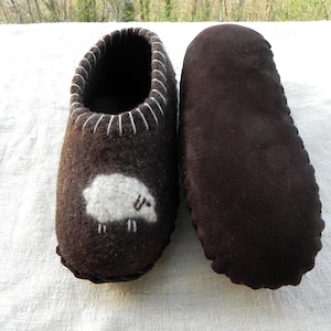 May include: A pair of brown felt slippers with a white sheep design on one slipper. The other slipper is plain brown.