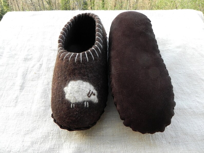 May include: A pair of brown felt slippers with a white sheep design on one slipper. The other slipper is plain brown.