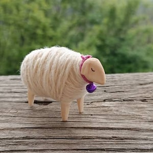 May include: A handcrafted wooden sheep figurine with a wool body and wooden legs and head. The sheep wears a pink collar with a purple bell. The figurine is set against a blurred green background.