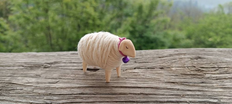 May include: A handcrafted wooden sheep figurine with a wool body and wooden legs and head. The sheep wears a pink collar with a purple bell. The figurine is set against a blurred green background.