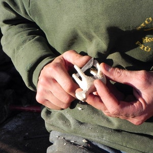 May include: A person carving a small, white wooden object with a knife. The object appears to be a stylized animal figure. The person is wearing a green long-sleeved shirt with text on the upper right side.