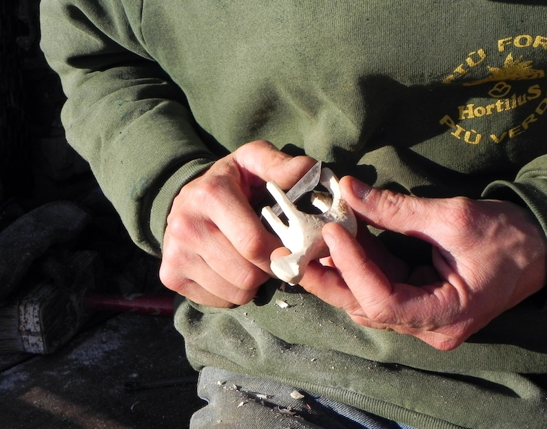 May include: A person carving a small, white wooden object with a knife. The object appears to be a stylized animal figure. The person is wearing a green long-sleeved shirt with text on the upper right side.