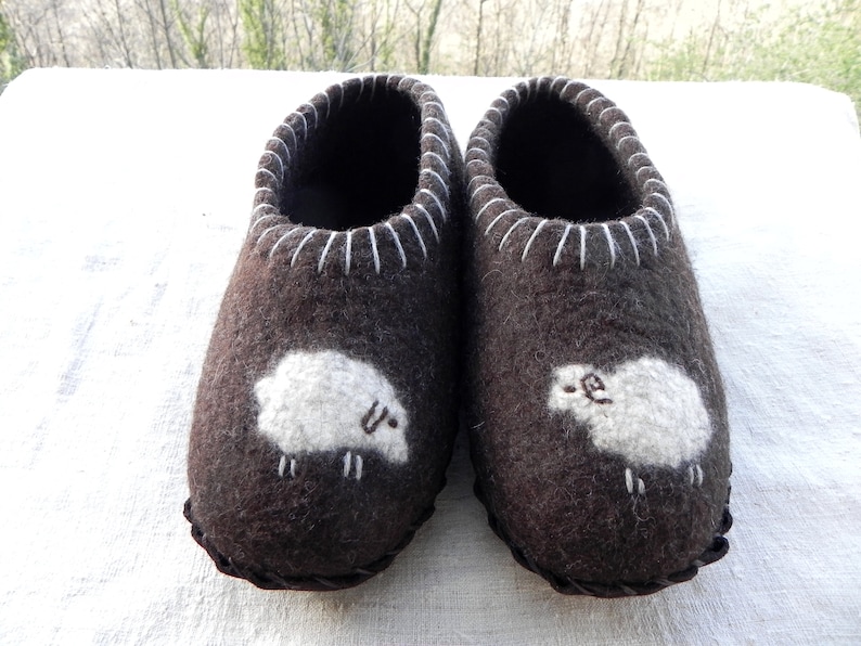 May include: A pair of brown wool slippers with white sheep embroidered on the front. The slippers have a white stitched trim around the top.