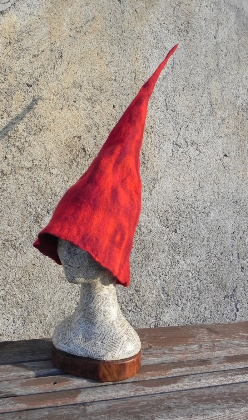 May include: A red felt conical hat with a pointed tip. The hat is displayed on a mannequin head with a newspaper-covered base.